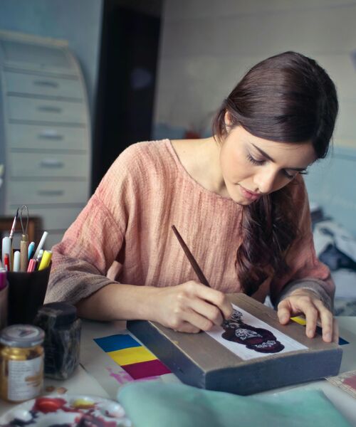 Eine Frau sitzt an einem Tisch und malt konzentriert an einem Kunstwerk, umgeben von Farben und Zeichenutensilien.
