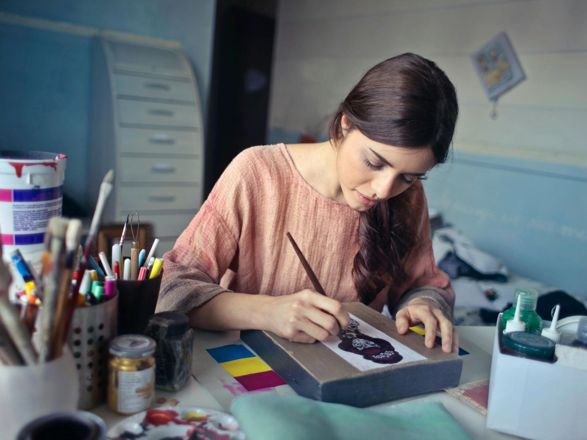 Eine Frau sitzt an einem Tisch und malt konzentriert an einem Kunstwerk, umgeben von Farben und Zeichenutensilien.