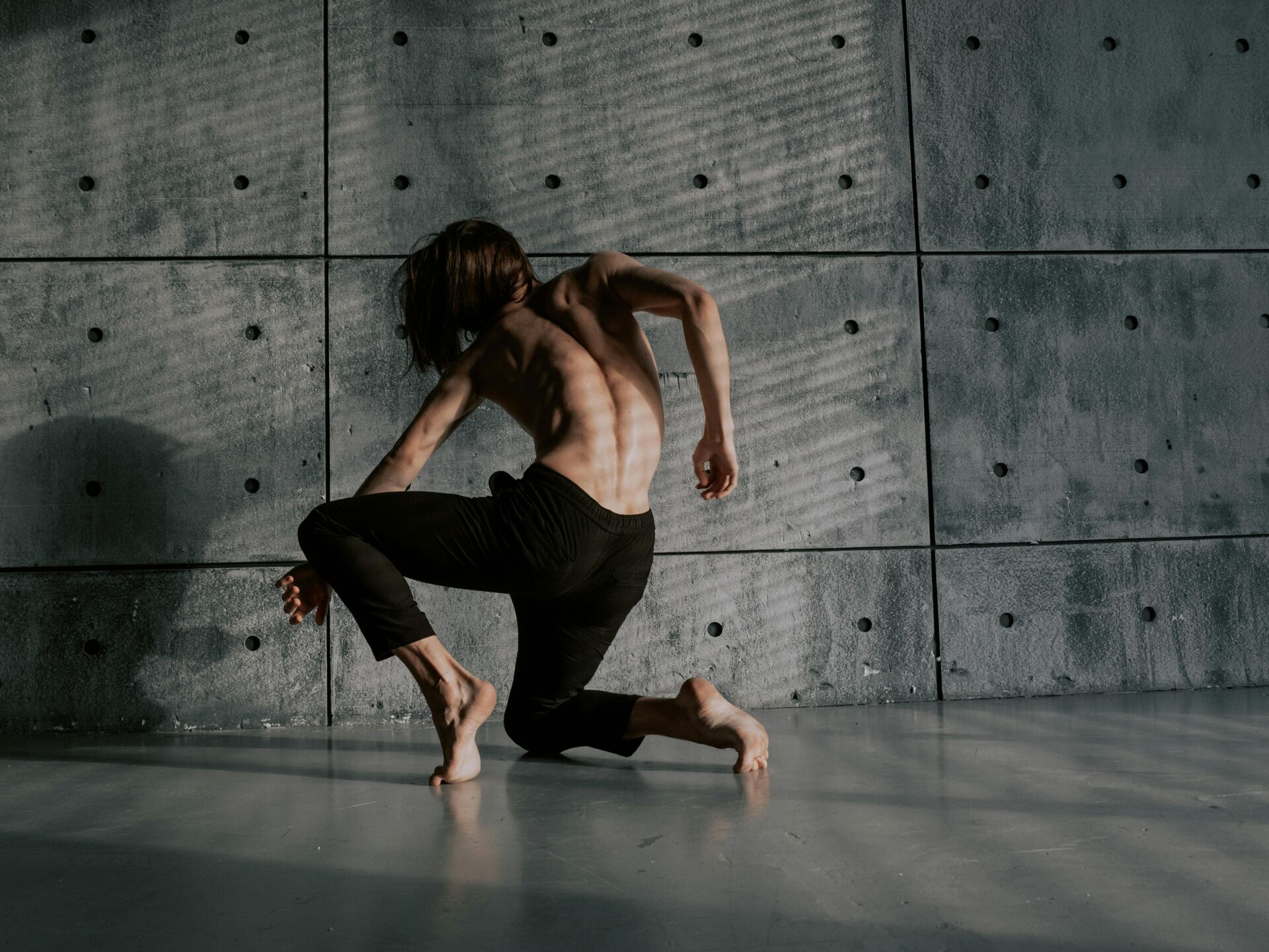 Eine Person mit nacktem Oberkörper bewegt sich tänzerisch auf dem Boden vor einer grauen Betonwand, das Licht wirft Schattenmuster auf ihren Körper.