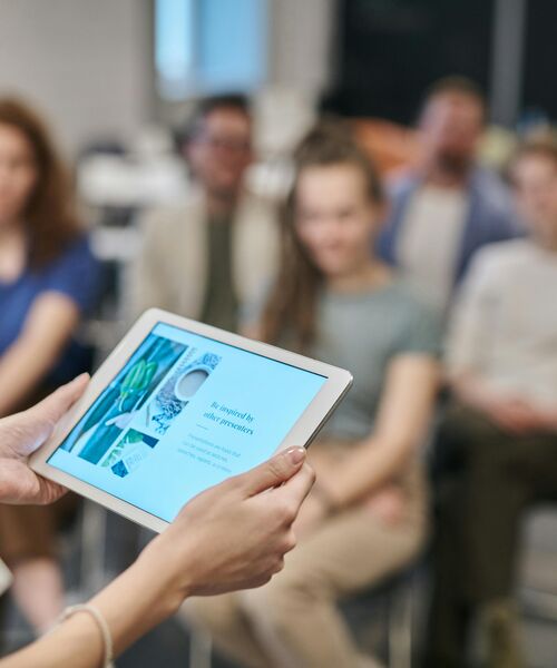Person hält ein Tablet mit einer Präsentation, im Hintergrund sitzt eine Gruppe von Menschen und hört zu.