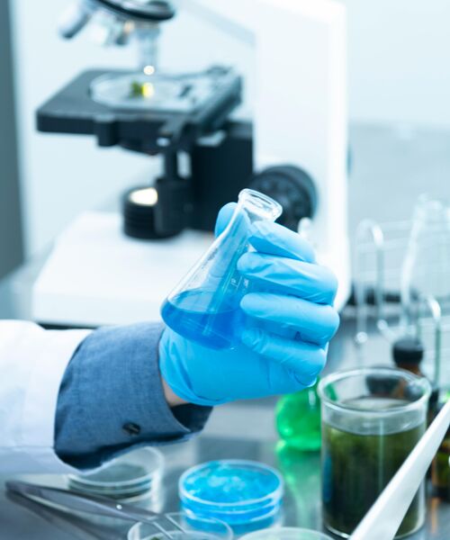 Laborumgebung mit einer Hand in blauen Handschuhen, die ein Becherglas mit blauer Flüssigkeit hält.