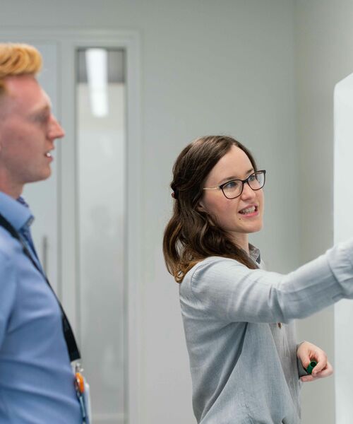 Frau erklärt etwas an einem Whiteboard, während ein Kollege aufmerksam danebensteht.