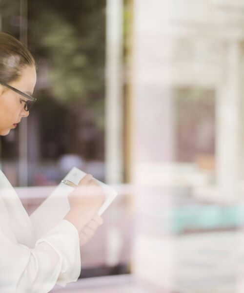 Frau schreibt konzentriert Notizen in ein Heft, im Hintergrund ein helles Büro mit Glasfront.